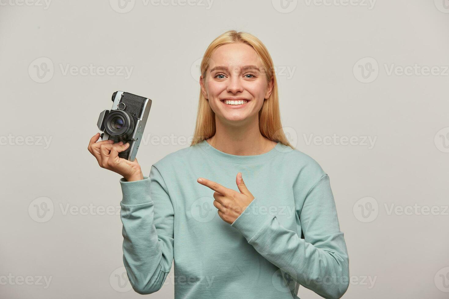 Lovely girl photographer taking a photo session, inspired by the retro vintage photo camera in hand, pointing on it with finger, dressed in blue sweatshirt, on grey background