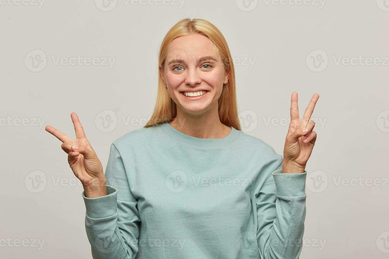 Lovely young blonde girl has fun, smiles, shows peace victory sign with fingers on both hands, wearing blue casual sweatshirt, isolated over grey background photo