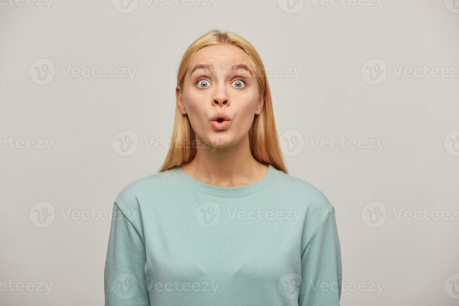Emotional blonde young woman, looking very surprised, open-eyed, whistles in surprise, wearing blue casual sweatshirt, see something unexpected in front. Human reaction concept over grey background photo