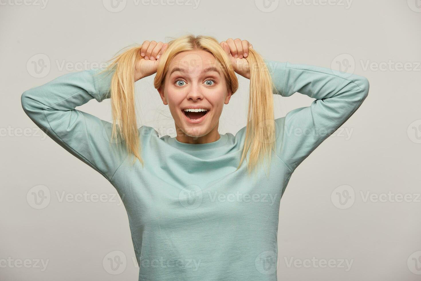 Lovely young blonde girl with long hair surprises, grimacing on camera, has fun jokes, rejoices, makes two tails on her head, wearing blue casual sweatshirt,isolated over grey background photo