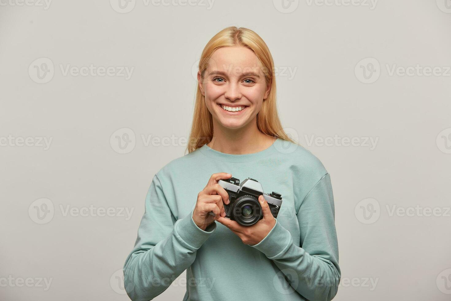 Blonde girl photographer, nicely widely smiles, looks happy delighted, holds in front a retro vintage photo camera in hands, dressed in blue sweatshirt, on grey background
