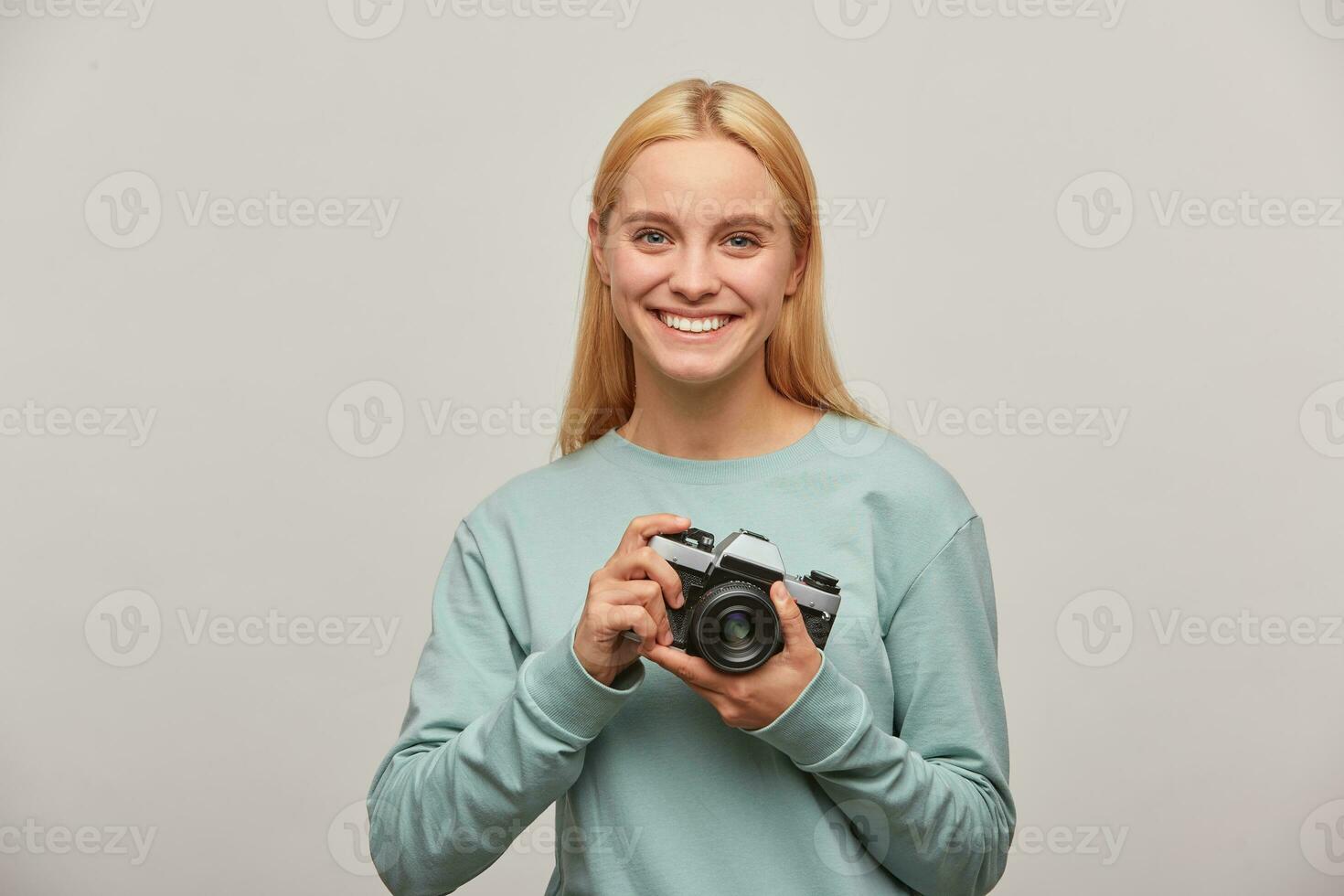 rubia niña fotógrafo, bien extensamente sonrisas, mira contento contento, sostiene en frente un retro Clásico foto cámara en manos, vestido en azul camisa de entrenamiento, en gris antecedentes