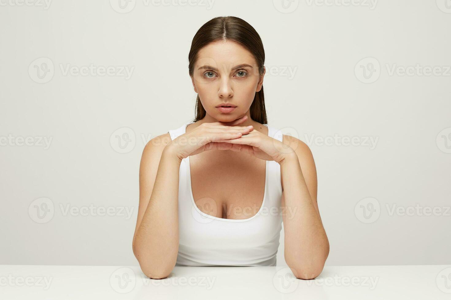 Beautiful woman with perfect healthy fresh skin sits at the table, looks at camera as in the mirror, palms folded under chin, carefully examines her face in details, not very satisfied with result photo
