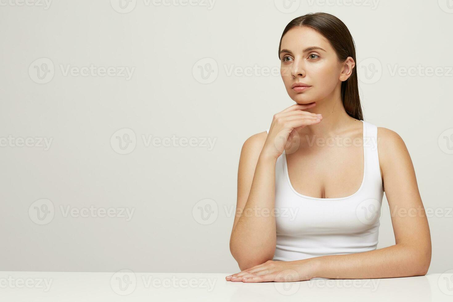 Attractive tender woman with perfect healthy fresh skin sits at the table, looks aside on a blank copy space, one hand is under the chin, hair pulled back, wears white T-shirt, over white background photo