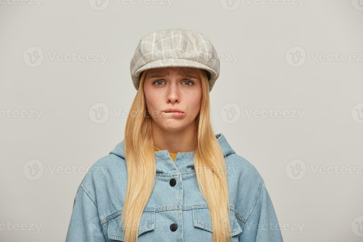 Lovely young blonde girl with long hair down, offended, upset, bit her lower lip, did not get what she wanted, wearing oversize denim jacket and beige checked hat cap,isolated over grey background photo