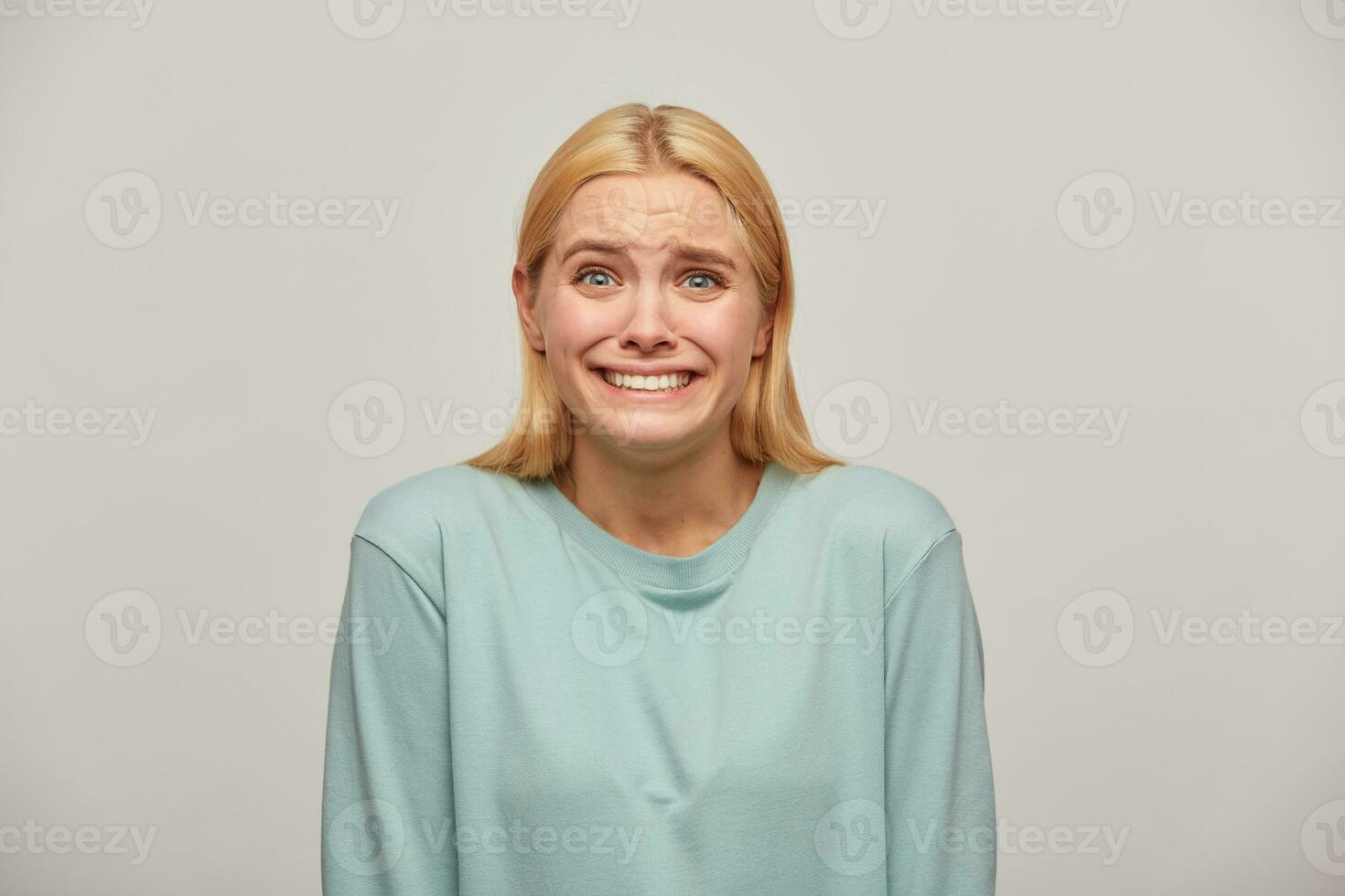 Scared blonde girl looks frightened afraid chattering teeth with fear, see something unexpected scary in front, here's to cry or scream, wearing blue casual sweatshirt,isolated over grey background photo