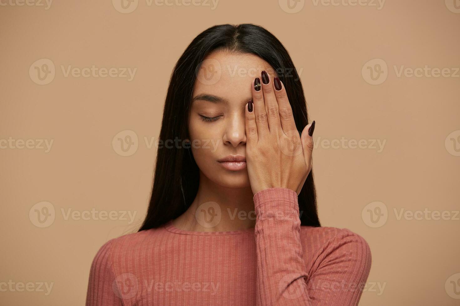 The pretty asian girl with her eyes closed hides half of her face with her palm, shows a beautiful manicure with sparkling pebbles, poses against beige background. photo