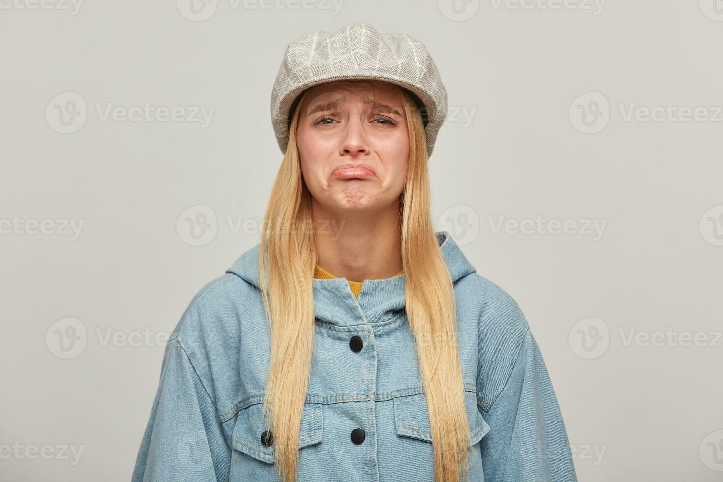 Emotional blonde girl, offended, upset, about to cry, lip pouting, did not get what she wanted, wearing oversize denim jacket and beige checked hat cap,isolated over grey background photo