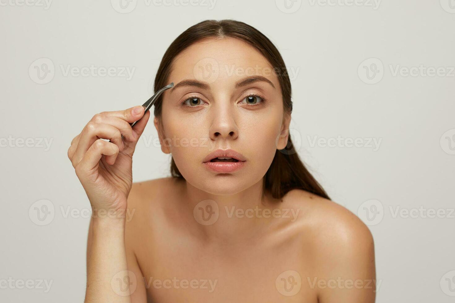 Close up of a beautiful woman as standing near a mirror. Young pretty girl cares for the face, plucks eyebrows with tweezers, isolated on white background. Head and shoulders photo