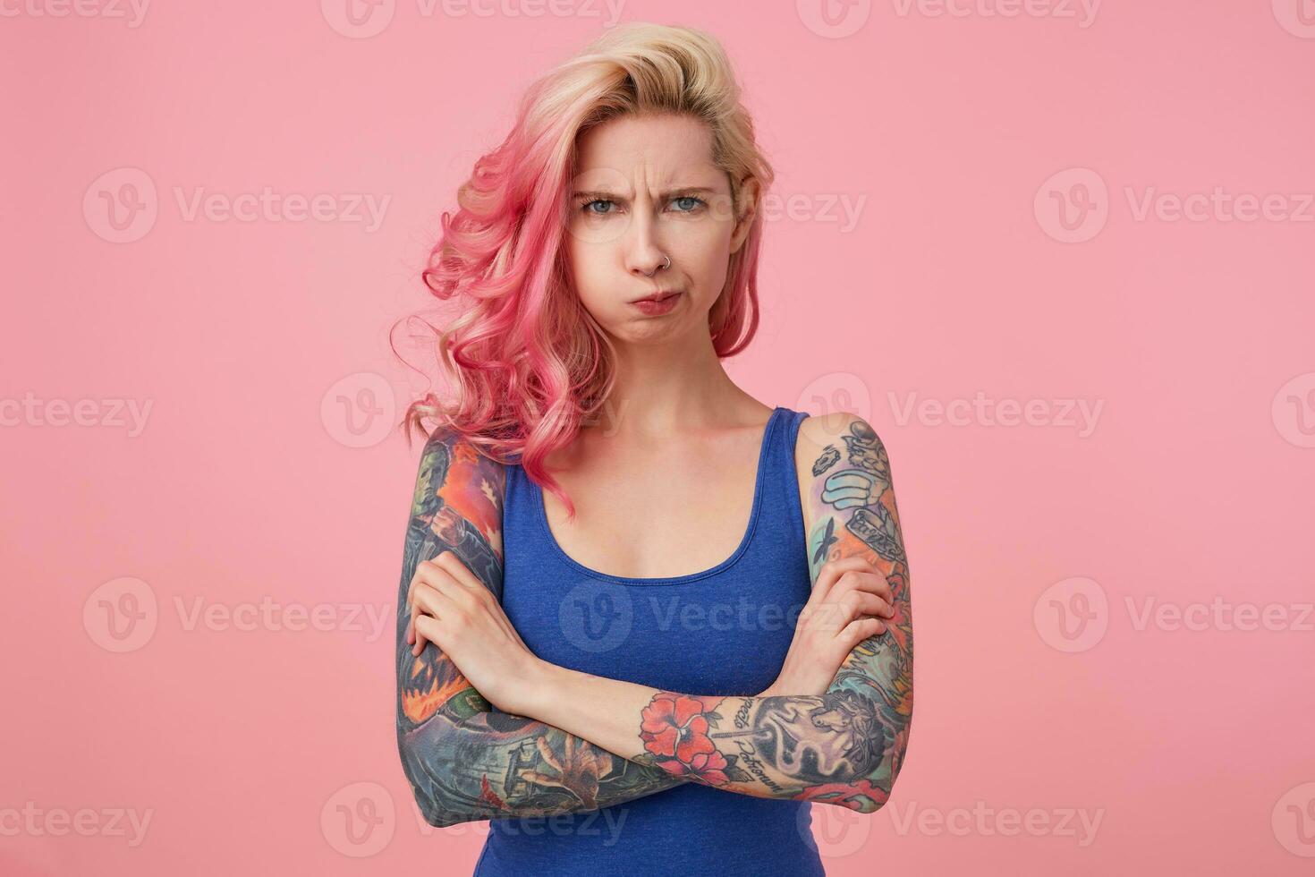 Young beautiful outraged pink haired woman with crossed arms, indignant looks at the camera, misunderstanding expression. Stands over pink background. photo
