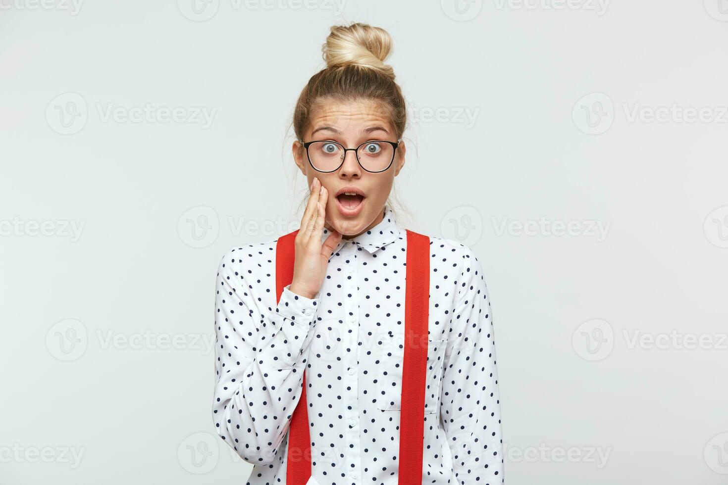 Young woman found out secret, gossip and spread rumors. Emotional girl feels amazed to hear confidential information. Dressed in a white shirt with black dots, glasses, red suspenders photo