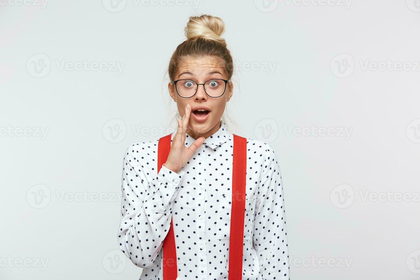 Young woman whispers secret, gossip and spread rumors. Emotional girl feels amazed to hear confidential information. Secrecy, gossiping. In a white shirt with black dots, glasses, red suspenders photo