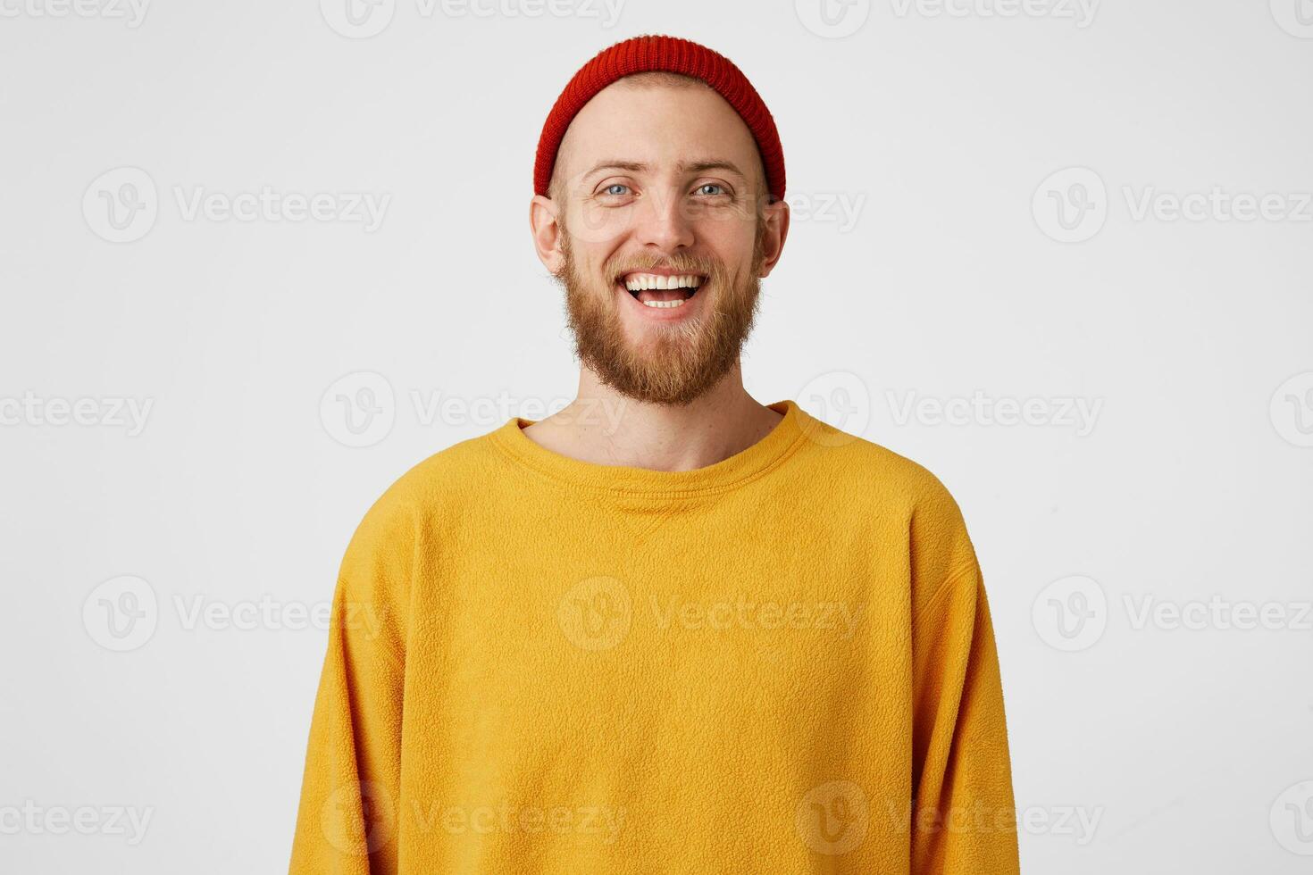 Isolated shot of joyful bearded young cute man laughs joyfully as hears funny joke, wears red hat and sweater, poses against white studio wall. Happiness and positive emotions concept photo