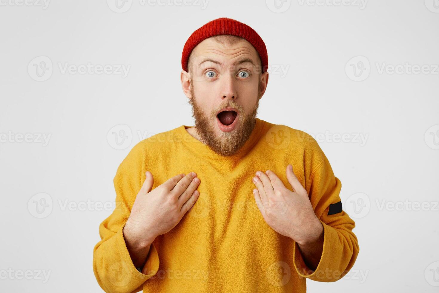 Amazed male keeps mouth opened, hands on breast, being shocked, stares at camera, isolated over white background. Bearded man in red hat man expresses great surprisement photo