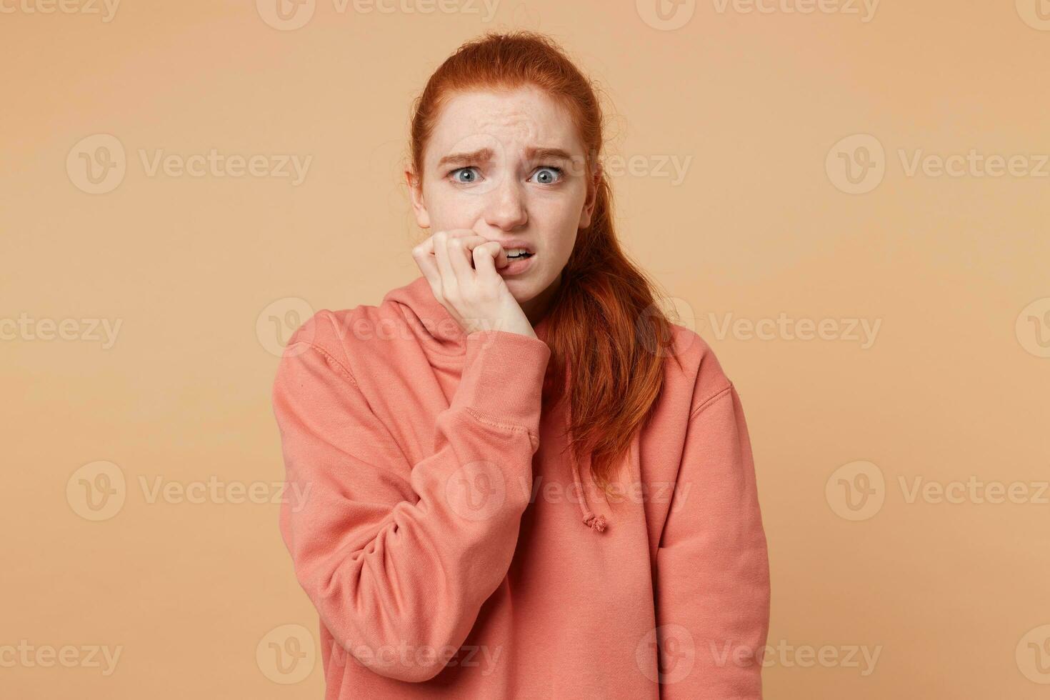 Portrait of a frightened young girl with blue eyes and red hair gathered in a ponytail that looks at the camera with fear,bites her nails, holds hand near the mouth,feels like a victim wants to escape photo