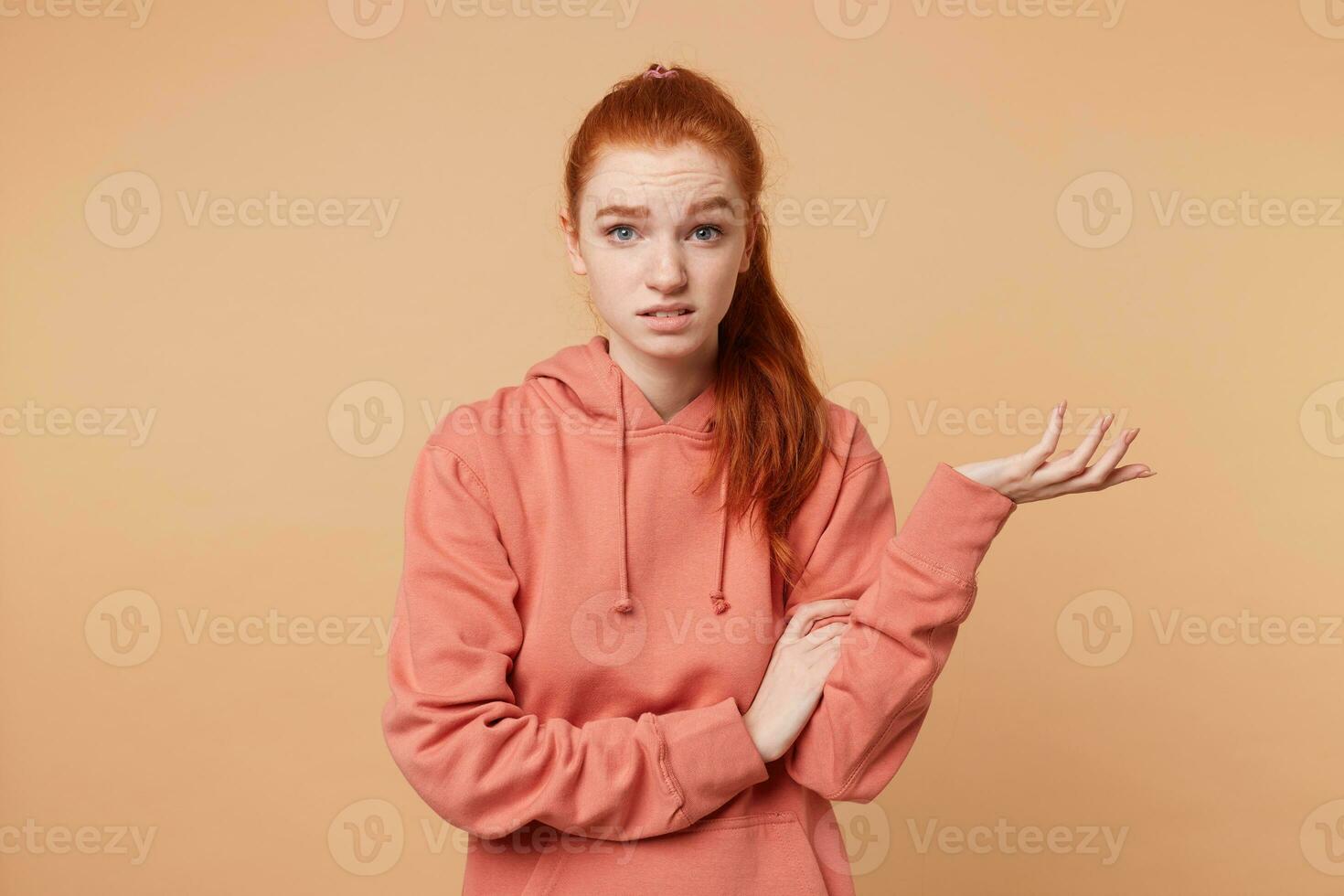 Dissatisfied red-haired girl with a tail dressed in hoodie with someone arguing one hand raised palm up trying to prove something, to defend their opinion, on beige studio background photo