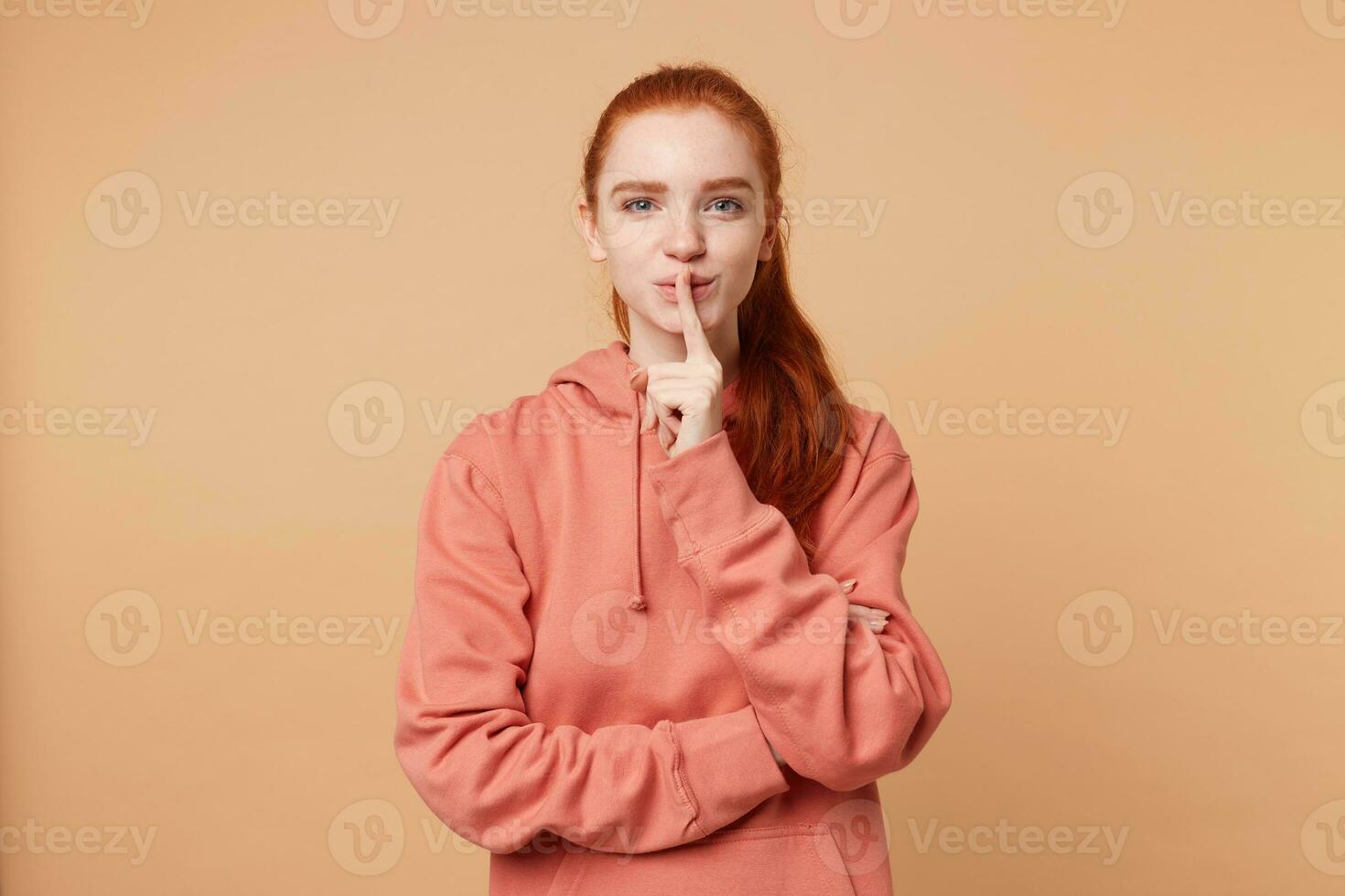 Cute red-haired girl with a hair gathered in ponytail nicely flirts, makes a silence gesture with index finger on her lips. Lets keep a secret, stay quiet, remain a privacy photo