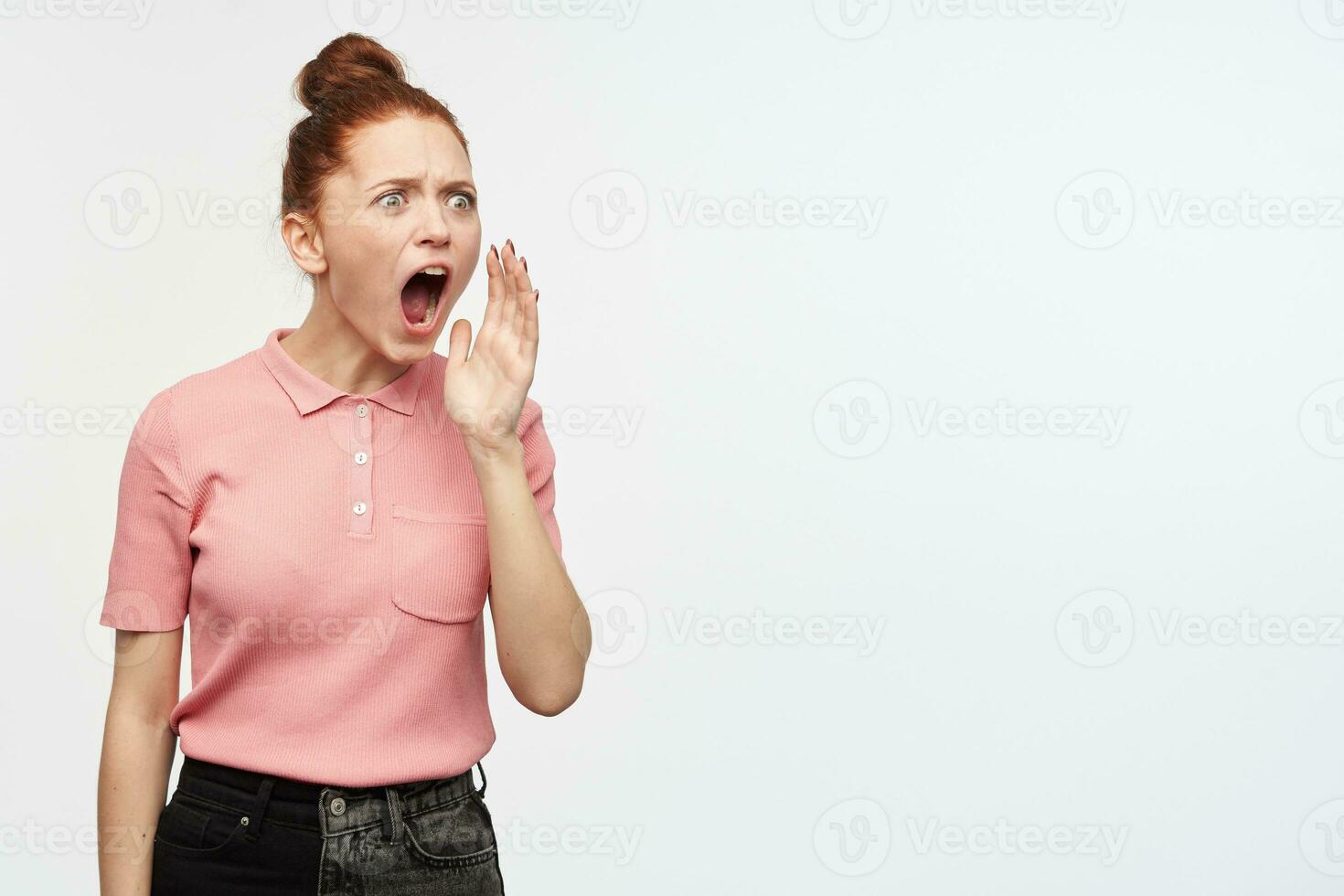 Portrait of shocked girl with ginger hair bun. Wearing pink t-shirt and black jeans. Shouts , holds palm next to mouth and watching to the right at copy space, isolated over white background photo