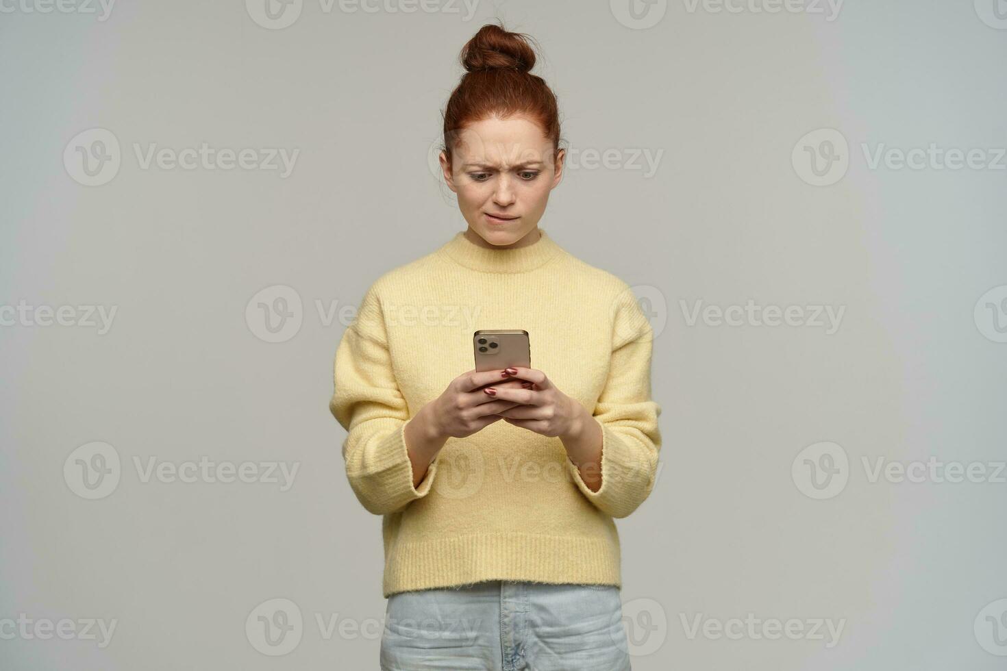 Young, focused woman with ginger hair bun. Wearing yellow sweater and holding a smartphone. Biting her lip and typing a message, watching at the phone. Stand isolated over grey background photo