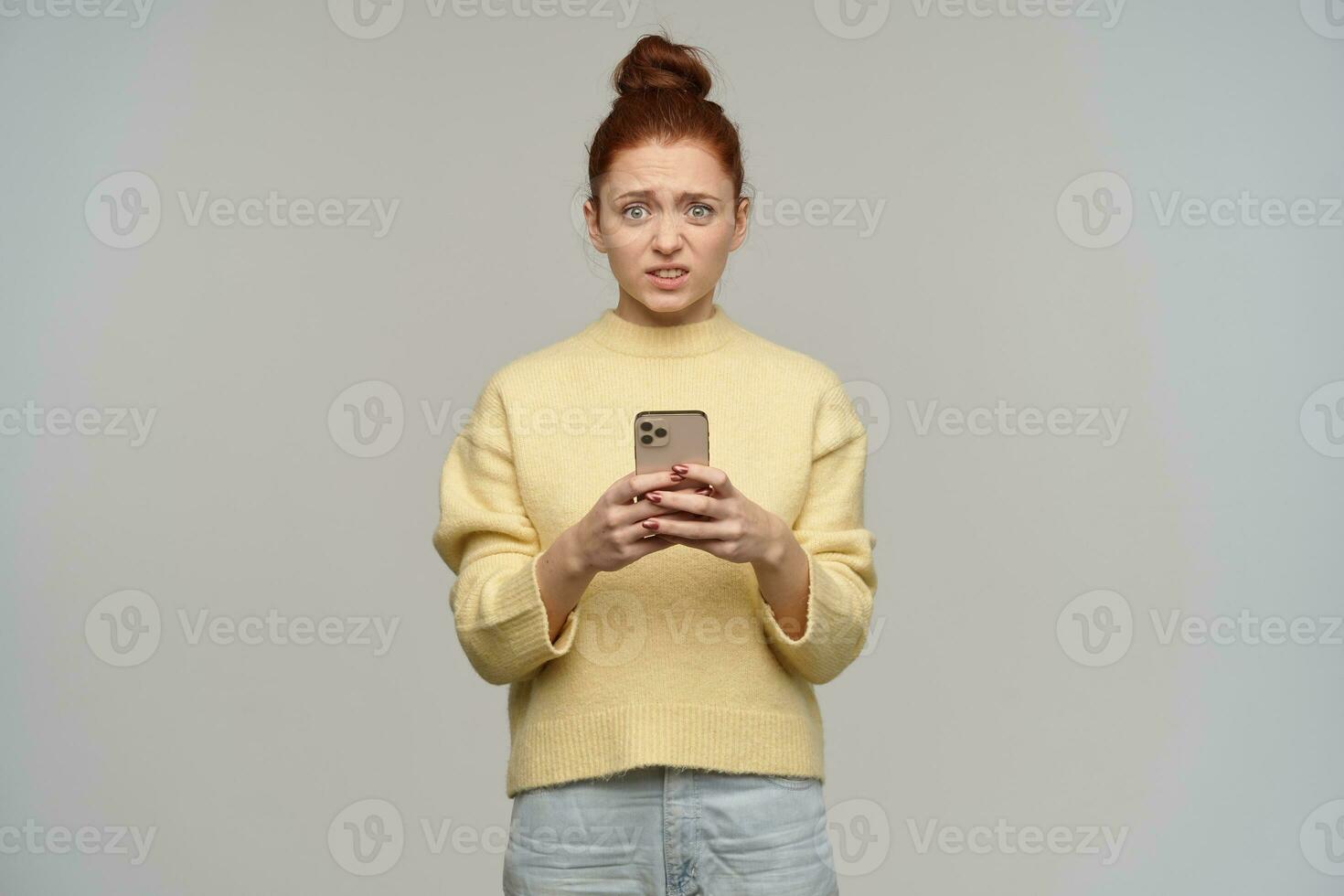 infeliz mirando mujer, disgustado pelirrojo niña con pelo bollo. vistiendo amarillo suéter y participación un teléfono inteligente tener visto alguna cosa negativo. acecho a el cámara, aislado terminado gris antecedentes foto