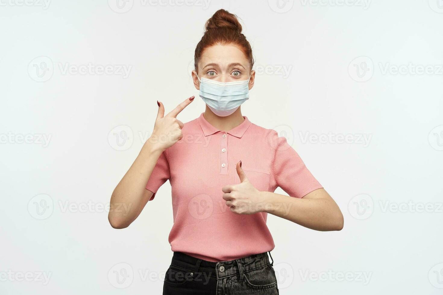 Portrait of girl with ginger hair bun. Wearing pink t-shirt and medical, protective face mask. Pointing at the mask and shows thumb up. Watching at the camera, isolated over white background photo