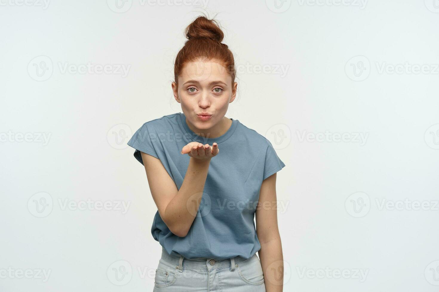 Positive, flirty woman with ginger hair bun. Wearing blue t-shirt. Sending air kiss, blow over her palm. Emotion concept. Watching lovely at the camera, isolated over white background photo