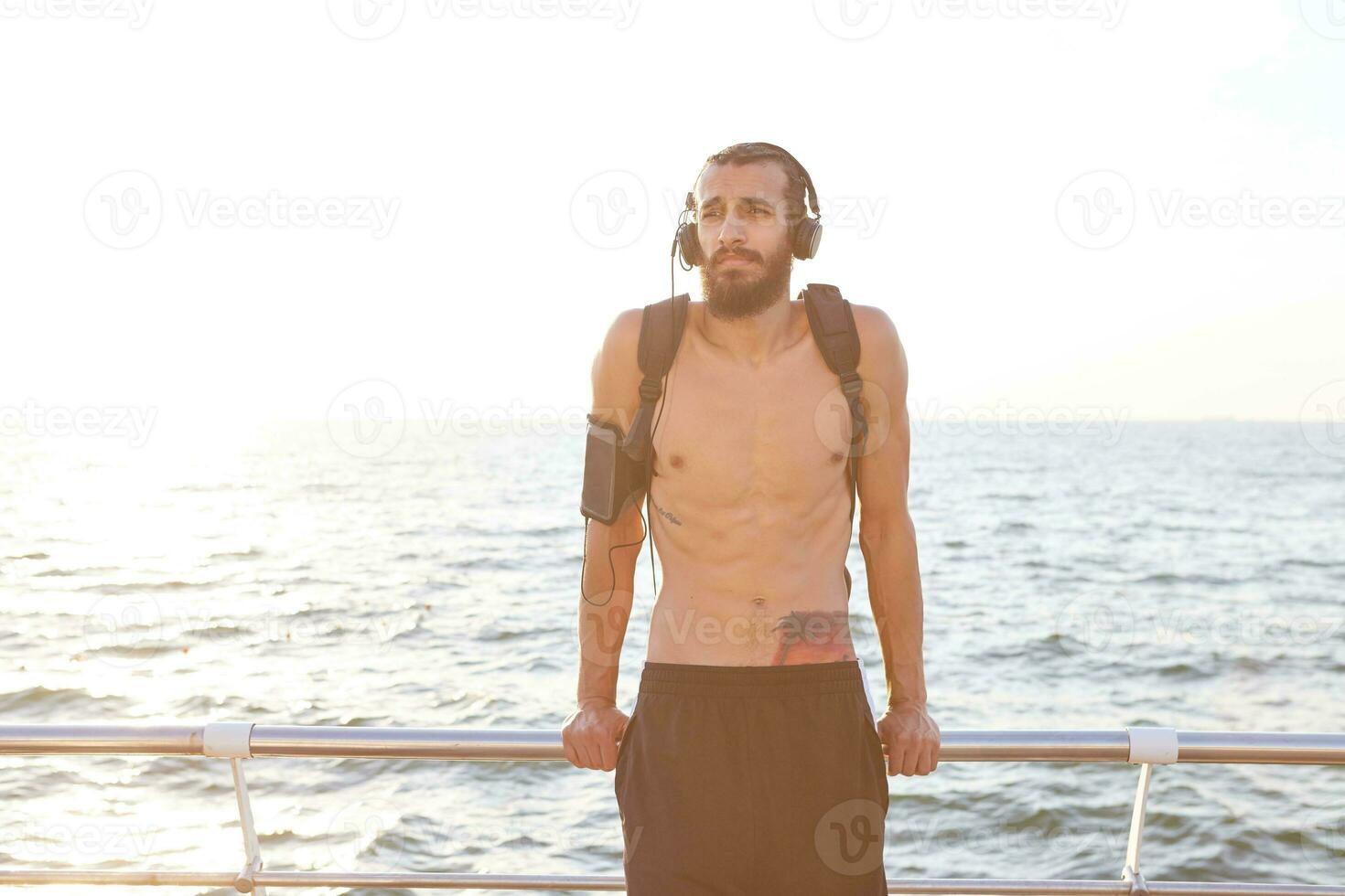 Young sporty guy with beard, rest after morning jogging, listen favorite mix on headphones at the seaside. photo