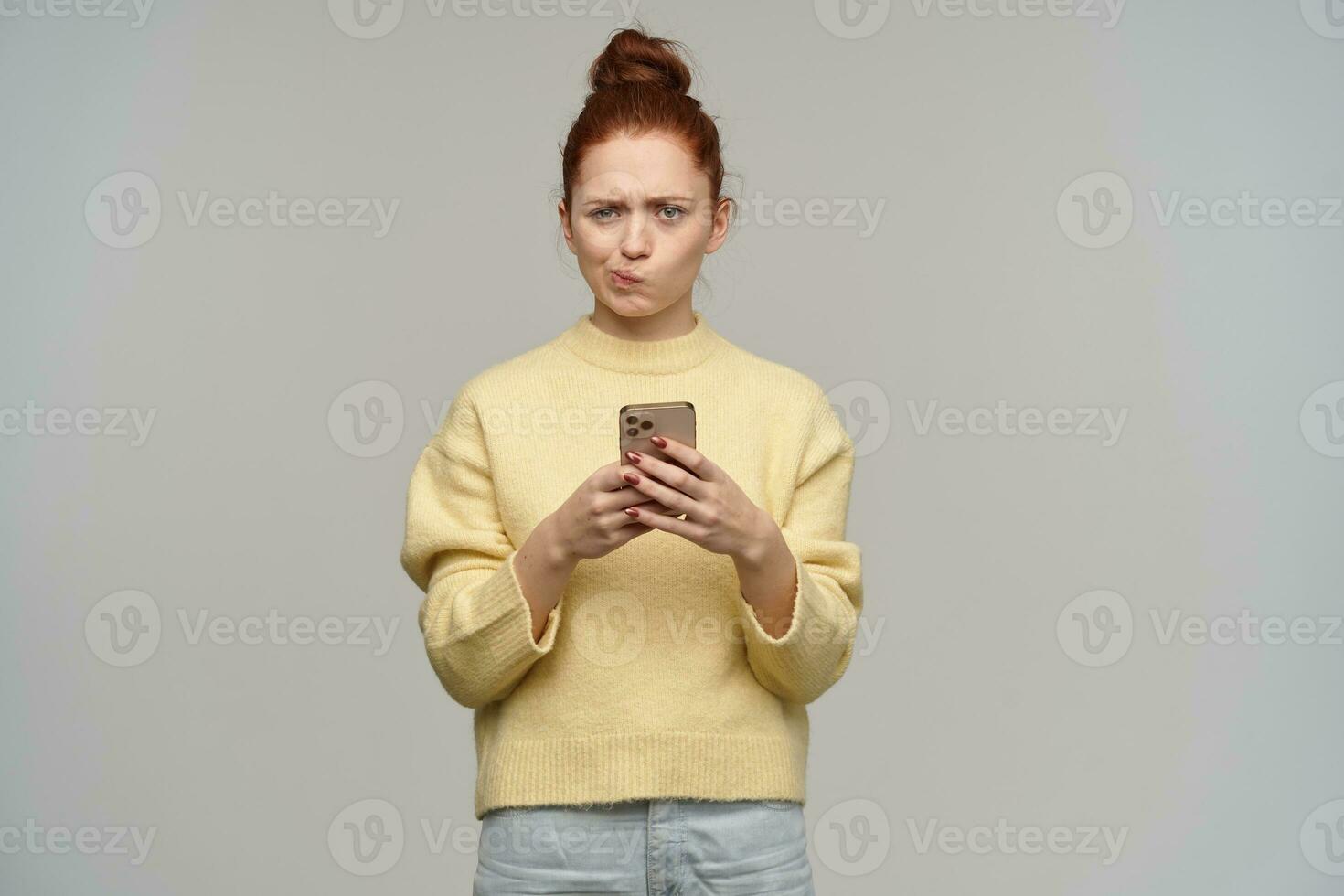 Frowning redhead girl with hair bun and pursed lips. Wearing yellow sweater and holding a smartphone. Having thoughts about content. Watching confused at the camera, isolated over grey background photo