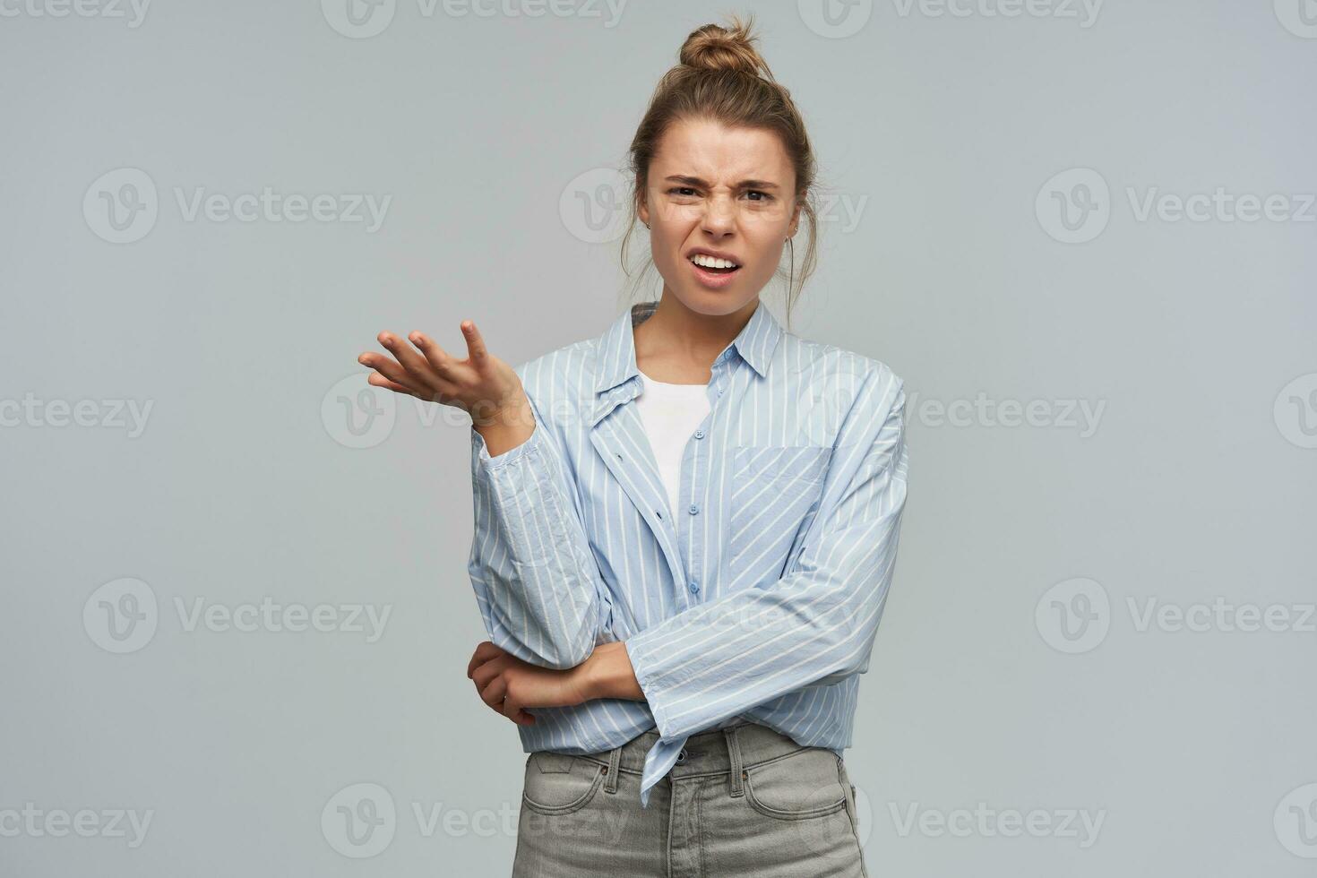 Confused, displeased woman with blond hair bun. Wearing blue striped knotted shirt. Emotion concept. Shrug her hand and frowns, asking. Watching unhappy at the camera, isolated over grey background photo