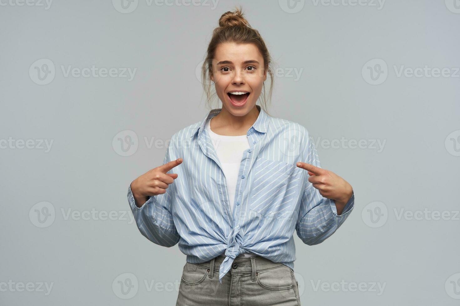 Young lady, pretty woman with blond hair bun. Wearing blue striped knotted shirt. Emotion concept. Pointing at herself, saying i'm the one. Watching at the camera, isolated over grey background photo