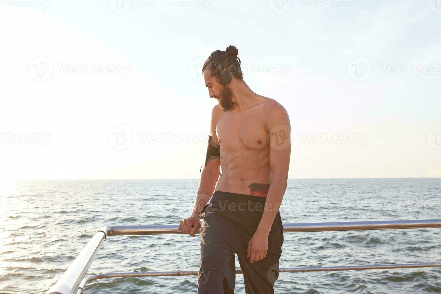 grandes Mañana retrato de joven deportivo hombre con barba, descanso después trotar a el playa, escucha favorito mezcla en auriculares. foto