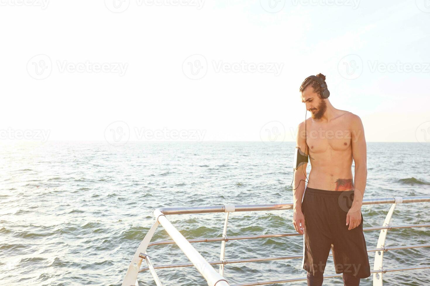 joven deportivo chico con barba, descanso después Mañana trotar a el playa, escucha favorito mezcla en auriculares. foto