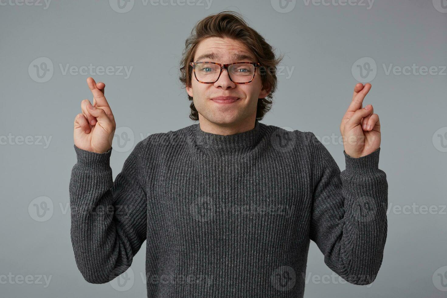 Photo of young handsome cheerful man with glasses wears in gray sweater, stands over gray background, crossed fingers and hopes for luck.
