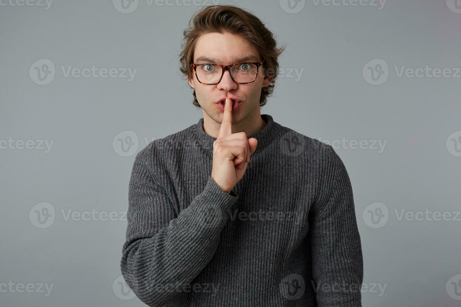 Make silence please Young man with glasses in gray sweater, shows silence sign, stands over gray background and keeps fore finger on lips, tries to keep conspiracy. photo