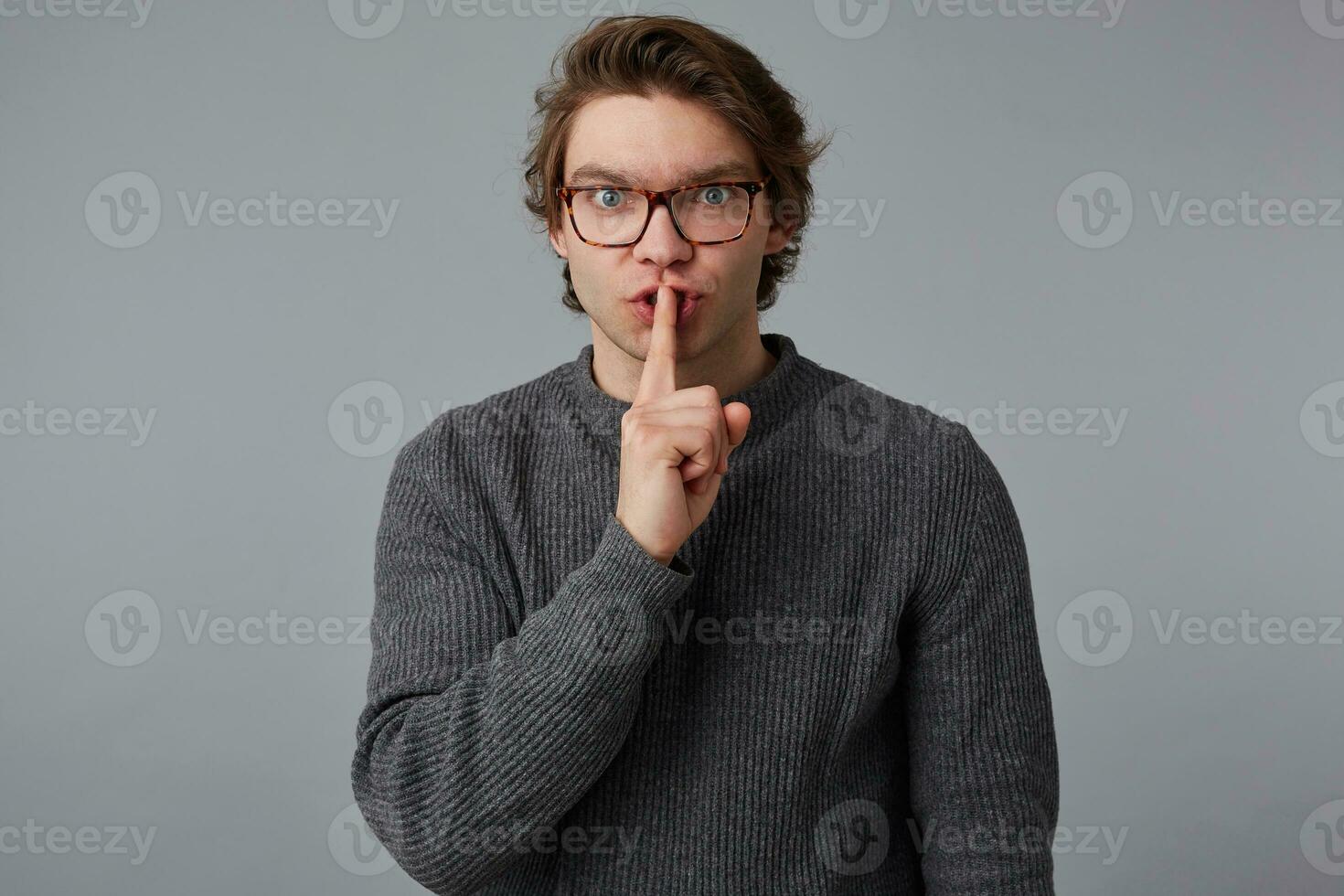 Make silence please Young man with glasses in gray sweater, shows silence sign, stands over gray background and keeps fore finger on lips, tries to keep conspiracy. photo