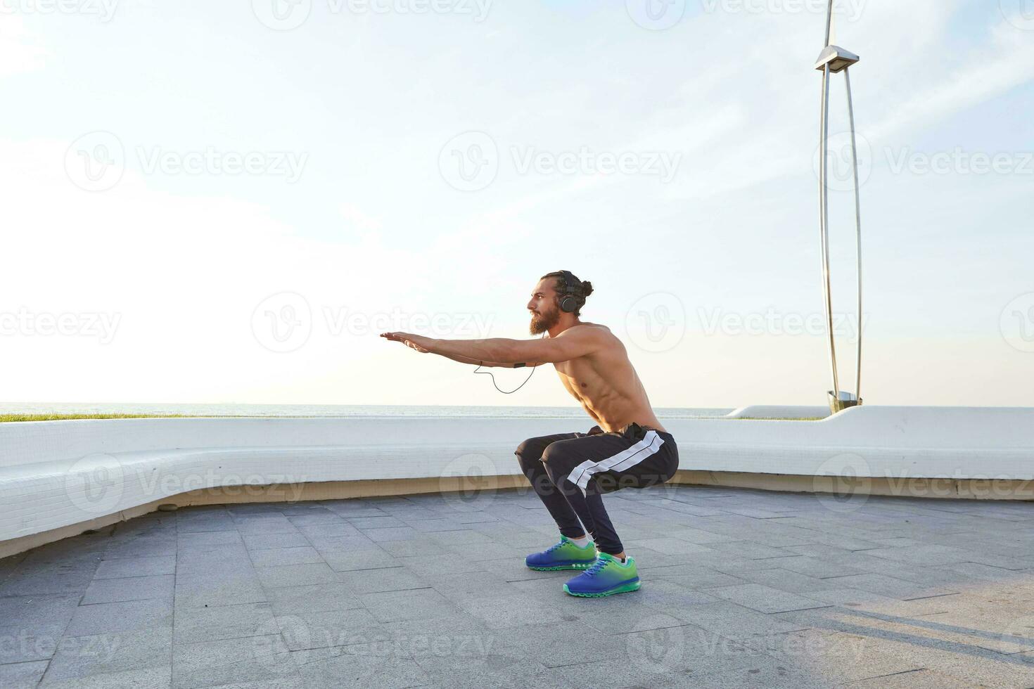 Bearded young man with fit athletic body, does exercises, listening music on headphones, has muscular body shape. photo