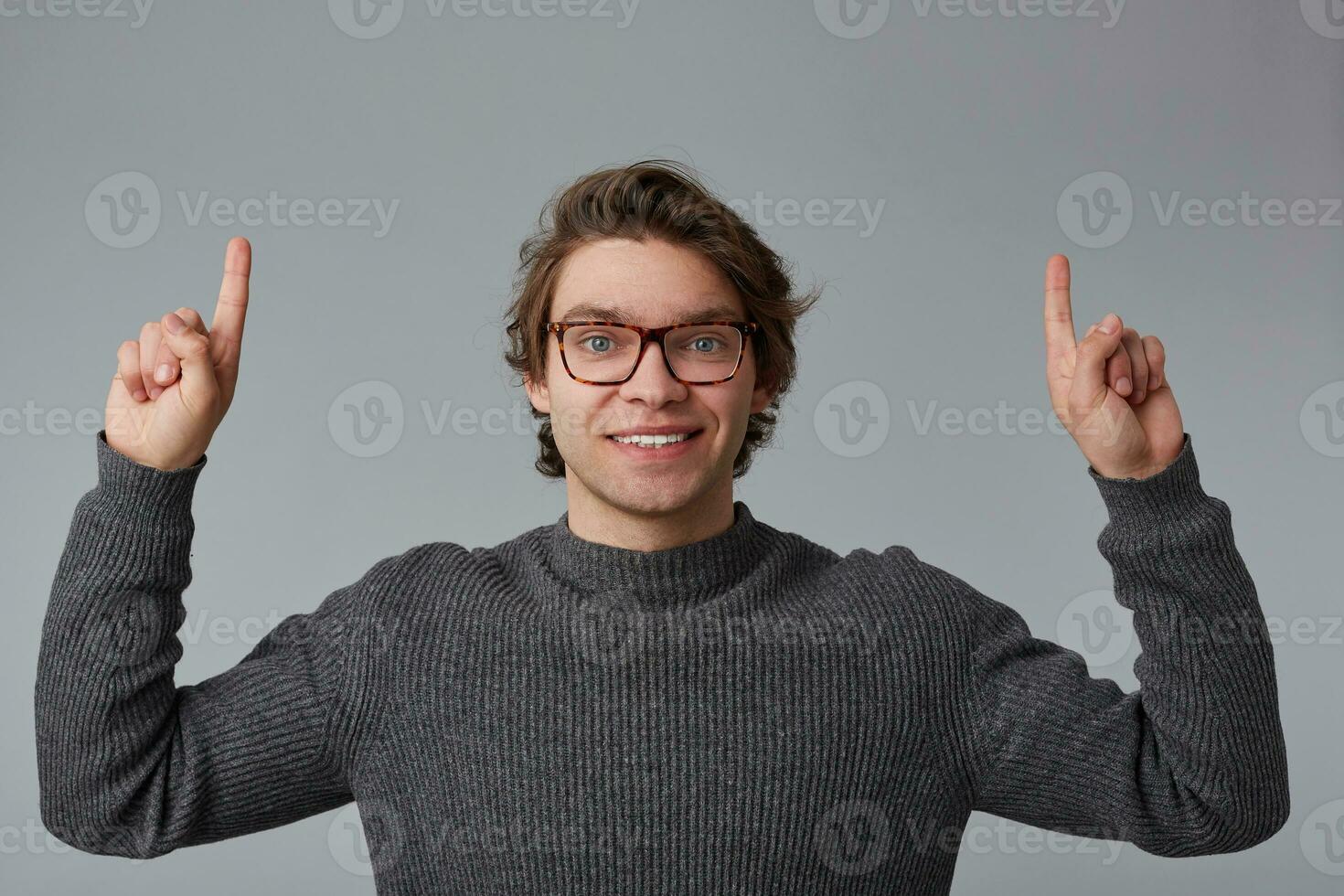 retrato de joven alegre hermoso hombre con lentes usa en gris suéter, soportes terminado gris antecedentes y en general sonrisas, puntos dedos arriba a un Copiar espacio terminado su cabeza. foto