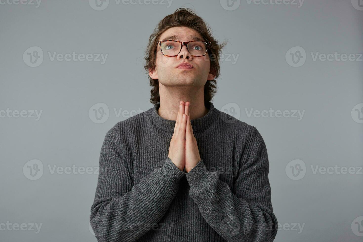 Portrait of young man with glasses wears in gray sweater, stands over gray background and looks up, has sorrorful expression, keeps palms in praying gesture, believes in something good. photo