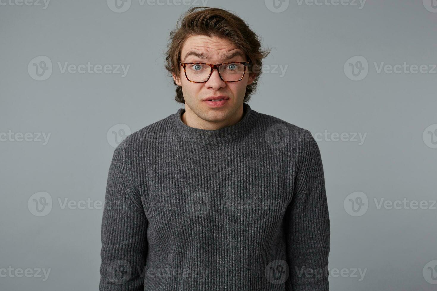 Portrait of young confused man with glasses wears in gray sweater, stands over gray background and looks at the camera with surprised expression. photo