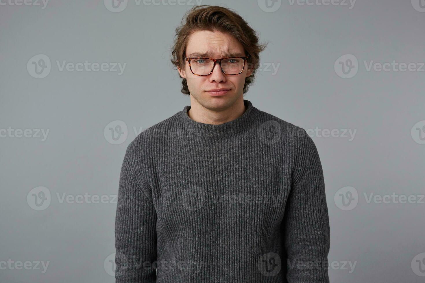 Photo of young sad handsome man with glasses wears in gray sweater, stands over gray background and looks unhappy.