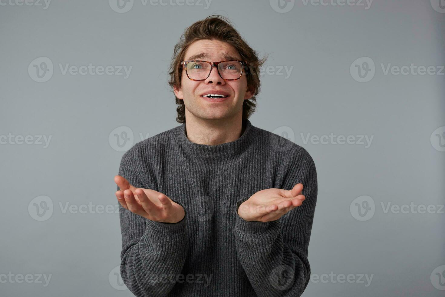 Young man with glasses wears in gray sweater, stands over gray background and looks up, has sorrorful expression, keeps palms up with praying expression. photo