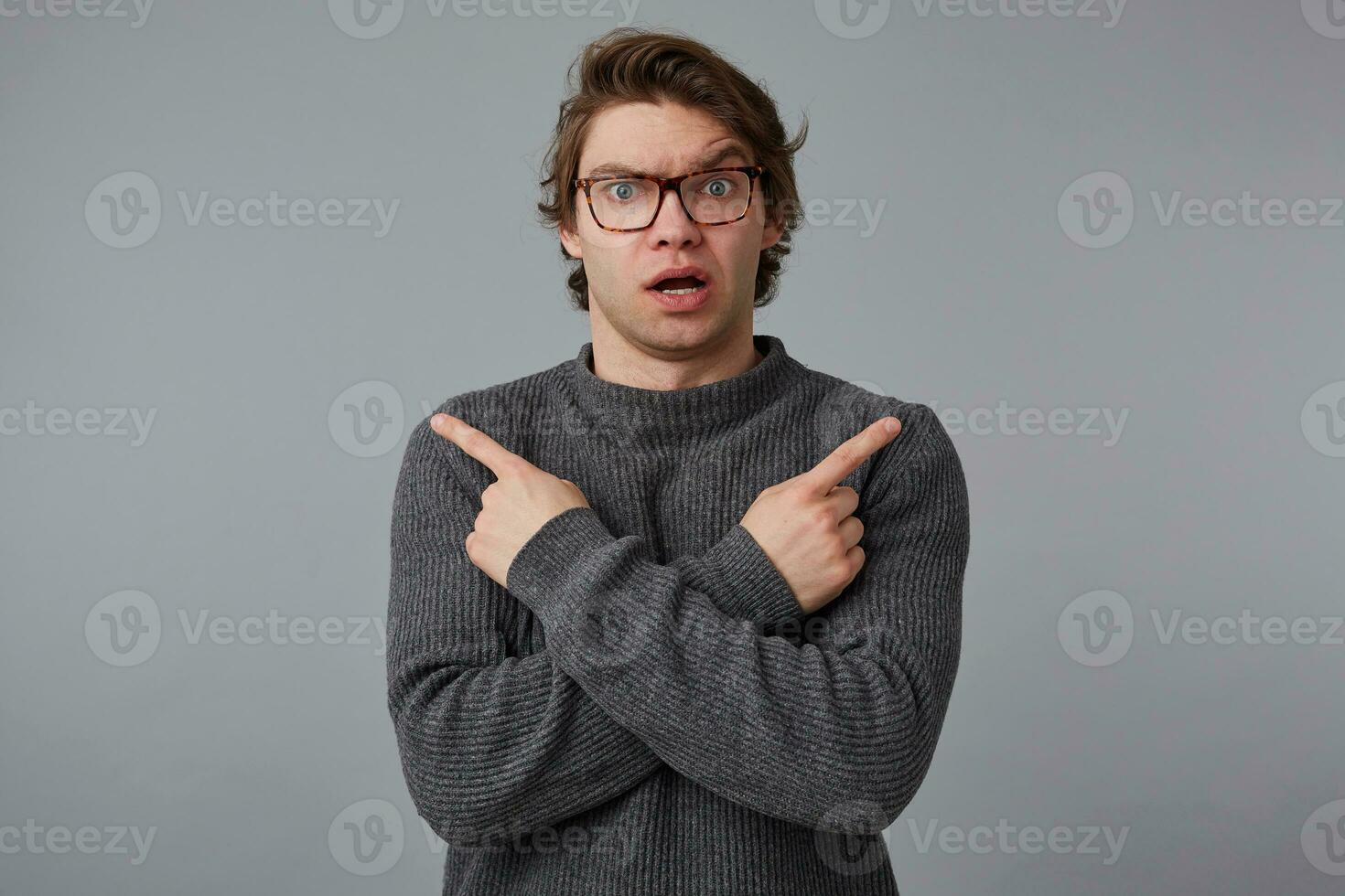 Portrait of young confused wondered man with glasses wears in gray sweater, stands over gray background, shows fingers in different directions. photo