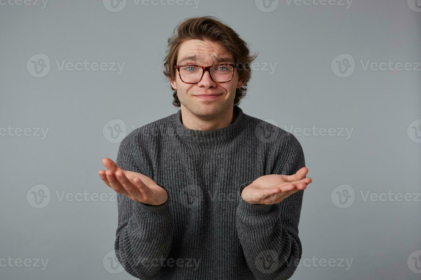 Photo of young pleaseful man with glasses wears in gray sweater, stands over gray background, and spreads his palms to the side.