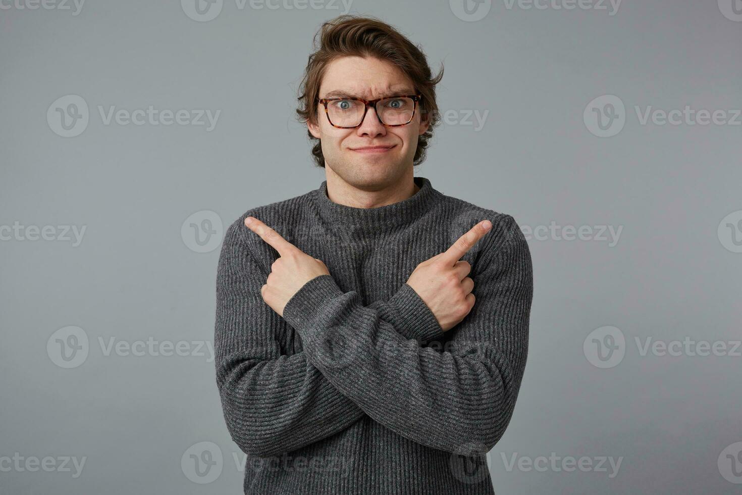 Photo of young confused wondered frowning man with glasses wears in gray sweater, stands over gray background, shows fingers in different directions.