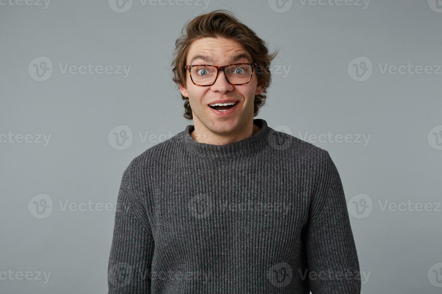 retrato de joven contento asombrado hombre con lentes usa en gris suéter, soportes terminado gris antecedentes con sorprendido expresión. foto