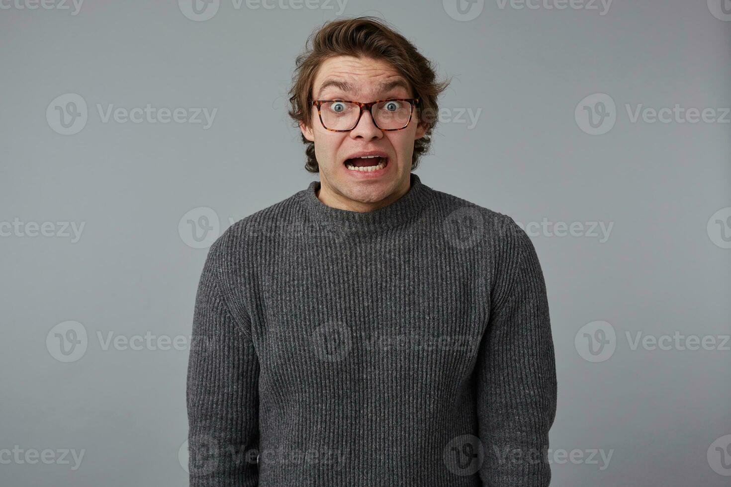 Portrait of young shocked guy with glasses, stands over gray background with wide open mouth and eyes, with surprised expression, looks dazed. photo