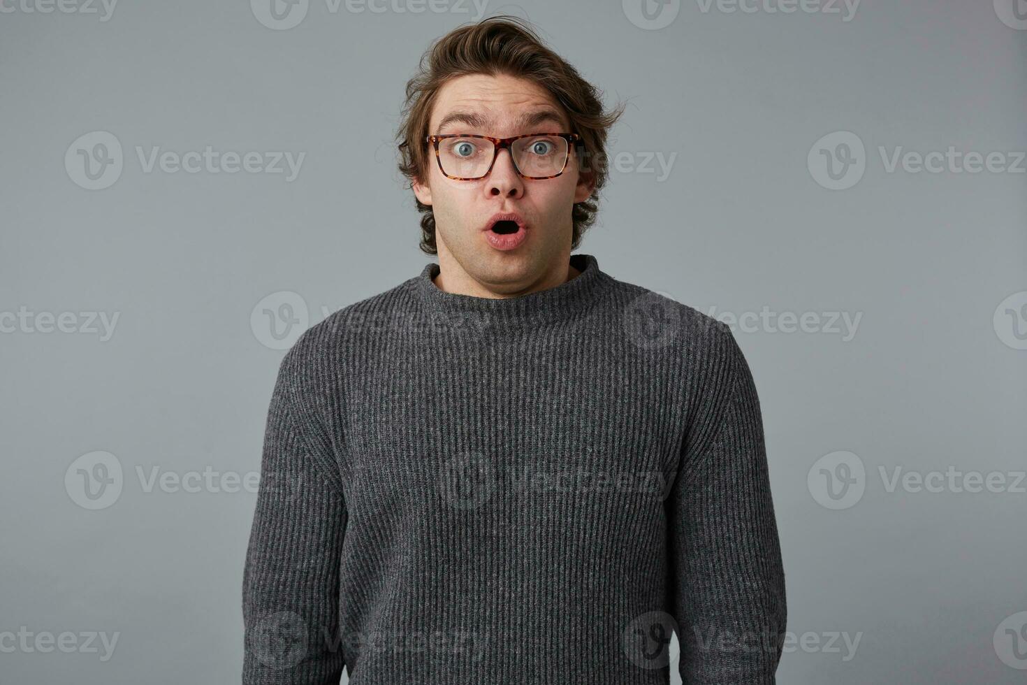 Portrait of young shocked man with glasses wears in gray sweater with wide open mouth and eyes, stands over gray background with surprised expression. photo