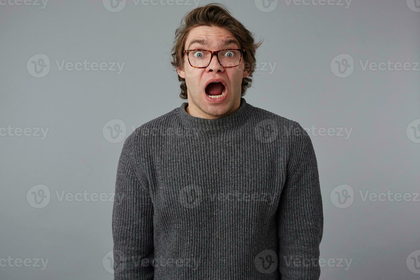 Portrait of young shocked guy with glasses, stands over gray background with wide open mouth and eyes, with surprised expression, looks scared. photo