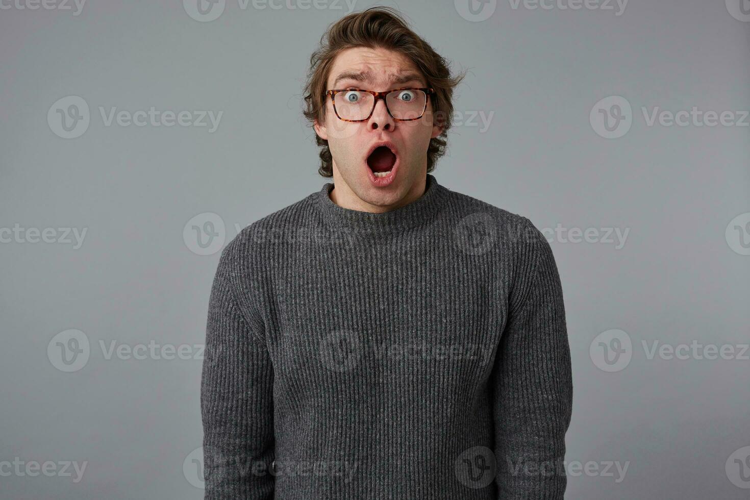 Portrait of young amazed guy with glasses, stands over gray background with wide open mouth and eyes, with surprised expression, looks scared and shocked. photo