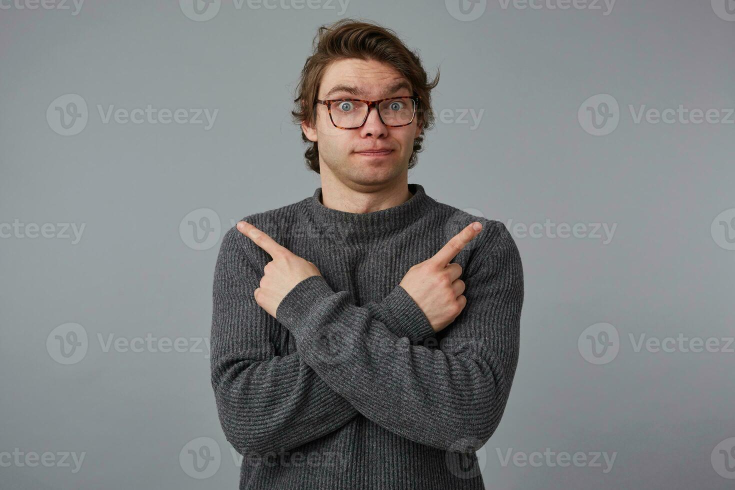 Photo of young confused pensive man with glasses wears in gray sweater, stands over gray background, shows fingers in different directions.